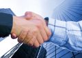 A close-up of a handshake between two businessmen with office buildings in the background